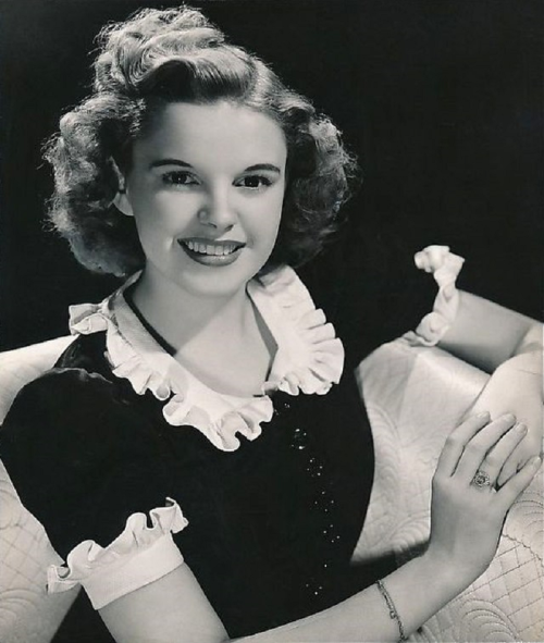 A vintage black-and-white portrait of a smiling young woman with curly hair, wearing a ruffled dress, seated elegantly.