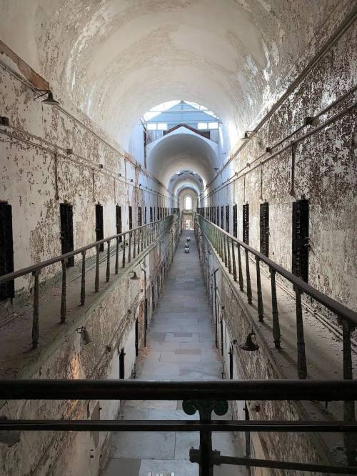 A long, empty prison corridor with peeling walls and barred windows, illuminated by natural light from above.