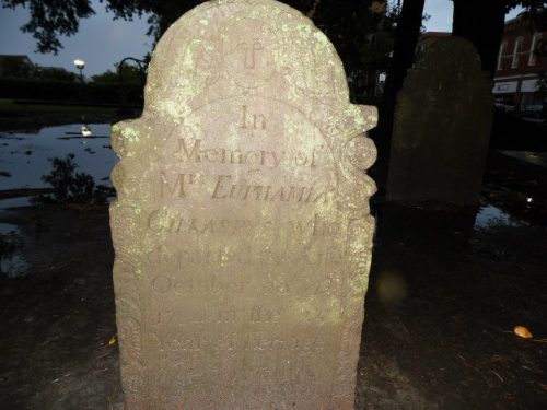 A weathered gravestone with inscriptions, partially obscured by water and foliage, in a cemetery setting.