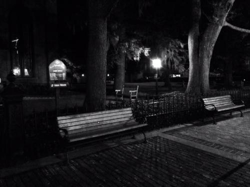 A dimly lit park scene at night featuring empty benches and trees, with a lamppost illuminating the area.