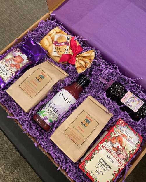 A gift box filled with snacks, including cookies, jam, and drink mixes, arranged on purple shredded paper.