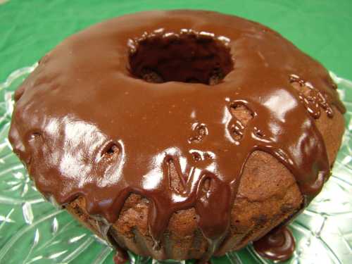 A chocolate-glazed bundt cake on a clear glass plate, with a glossy, dripping chocolate topping.