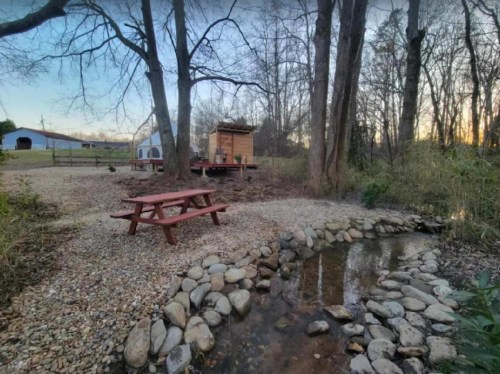 A serene outdoor scene featuring a picnic table, a small stream, and a wooden shed surrounded by trees at sunset.