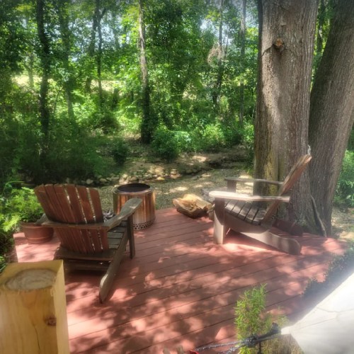 A cozy wooden deck in a forested area, featuring two chairs, a fire pit, and lush greenery surrounding it.