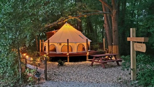A cozy glamping tent illuminated at night, surrounded by trees and a picnic table nearby.