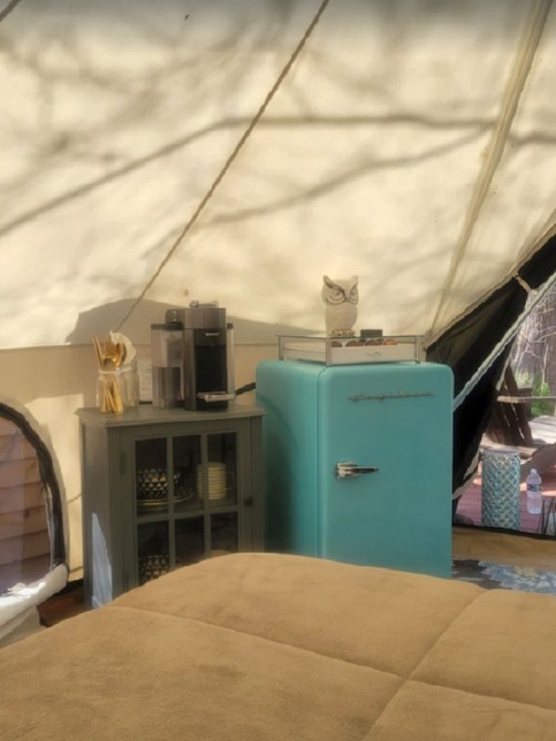 Interior of a tent featuring a vintage blue fridge, a coffee maker, and a cozy bed with soft bedding.
