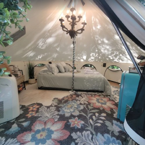 Cozy glamping tent interior with a bed, chandelier, and floral rug, surrounded by greenery and soft lighting.