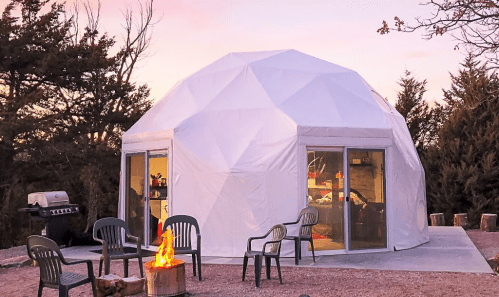A geodesic dome structure with large windows, surrounded by chairs and a fire pit, set in a natural landscape at sunset.