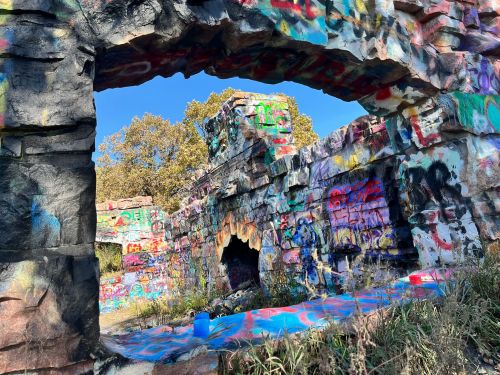 Graffiti-covered stone ruins with an archway, surrounded by greenery and a clear blue sky.