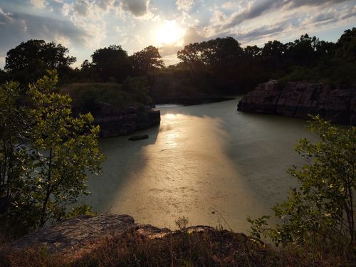 Sunset over a tranquil pond surrounded by rocky cliffs and lush greenery, with reflections on the water's surface.