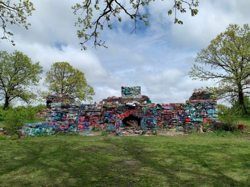 Colorful graffiti covers the ruins of a stone structure, surrounded by green grass and trees under a cloudy sky.