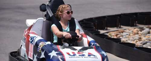 A young girl drives a go-kart decorated with the USA flag design on a racetrack.