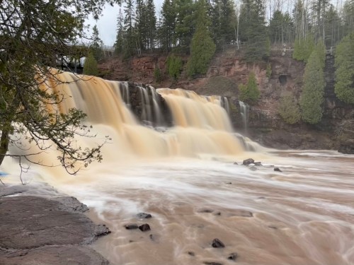 A cascading waterfall flows over rocky terrain, surrounded by lush green trees and a cloudy sky.