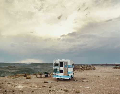 A vintage camper trailer sits alone in a vast, arid landscape under a cloudy sky.