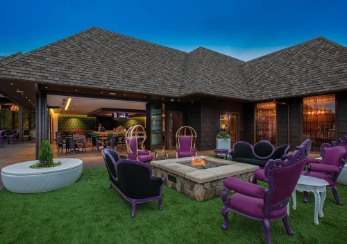 Outdoor lounge area with purple chairs, a stone fire pit, and a modern bar in the background, surrounded by greenery.