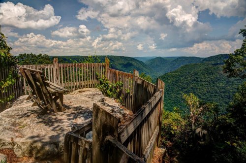 A scenic overlook with a wooden fence, featuring a chair on a rocky outcrop, surrounded by lush green mountains and clouds.