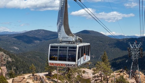 A gondola lifts over a mountainous landscape, with trees and distant peaks under a blue sky.