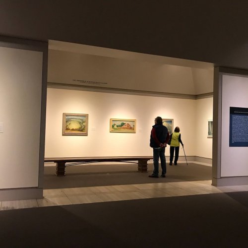 Two visitors observe paintings in a gallery, with one person standing and the other seated on a bench.