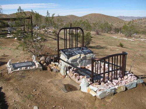 A unique grave marked by a bed frame and stones, set in a desert landscape with hills in the background.