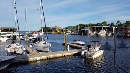A marina scene with several boats docked, people walking on the pier, and buildings in the background under a blue sky.