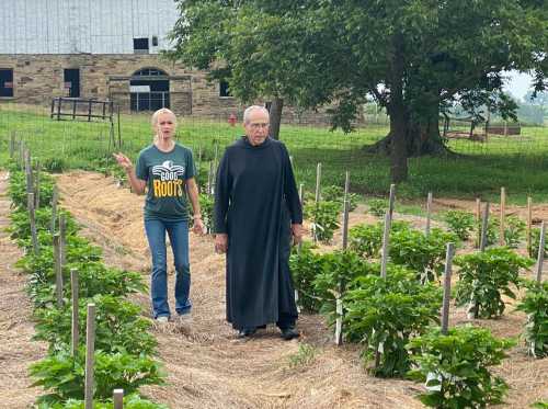 A woman in a t-shirt walks alongside a man in a robe through a garden with rows of plants.