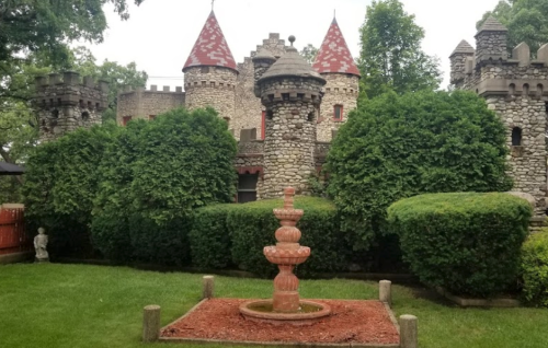 A stone castle with red-tipped towers surrounded by lush greenery and a small fountain in the foreground.