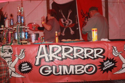 A festive booth with a pirate-themed banner reading "ARRRRRR GUMBO," showcasing trophies and people preparing food.