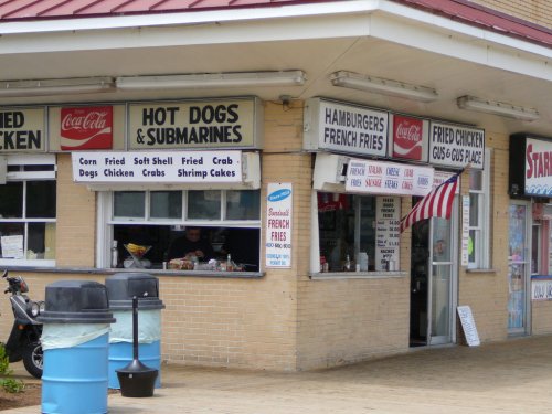 A food stand with signs for hot dogs, hamburgers, and fried chicken, featuring a man working behind the counter.