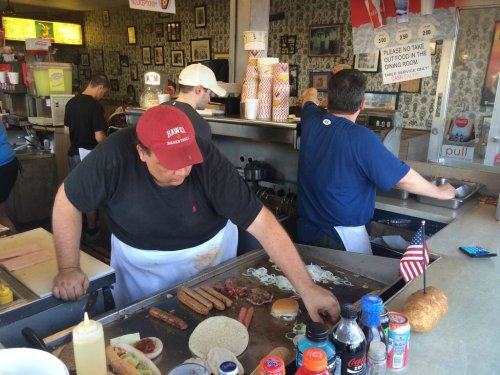 Two cooks prepare food at a busy diner, with various ingredients and condiments on the counter. American flags are visible.