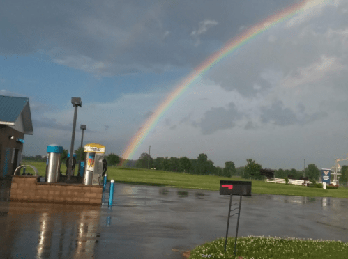 A vibrant rainbow arcs over a gas station after rain, with wet pavement reflecting the colorful sky.