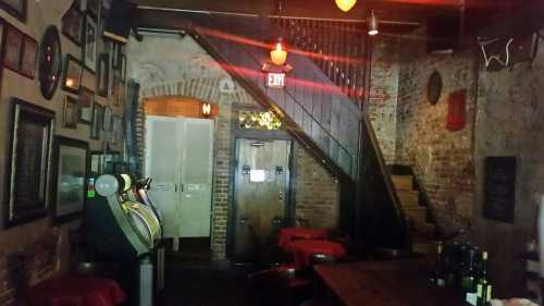 Dimly lit bar interior with exposed brick walls, a jukebox, and a staircase leading to an upper level.