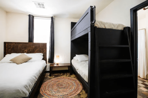Cozy bedroom featuring a bunk bed, a double bed, a nightstand, and a colorful rug. Natural light from a window.