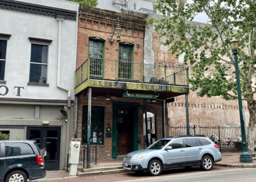 Historic brick building with a balcony, surrounded by trees and parked cars in an urban setting.