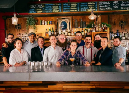 A diverse group of bartenders and staff pose together behind a bar, showcasing cocktails and a lively atmosphere.