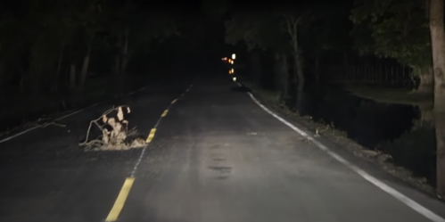 A dark, empty road at night with a small pile of debris on the pavement and trees lining the sides.