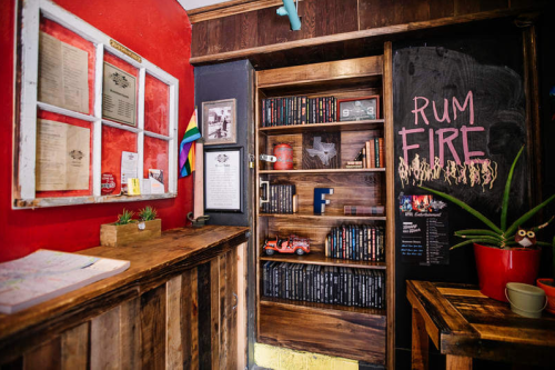 Cozy interior of a bar with wooden decor, a bookshelf, and a chalkboard displaying "RUM FIRE."