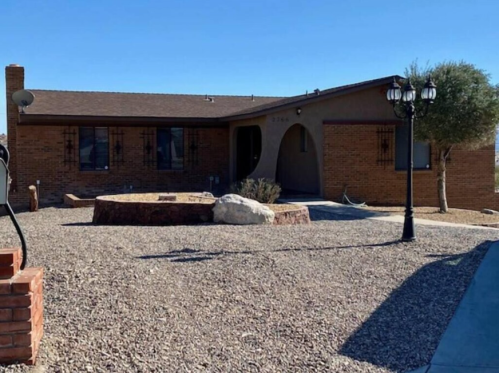 A single-story brick house with a gravel yard, featuring a circular stone fire pit and decorative lamp posts.