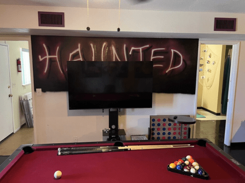 A pool table in a room with a TV and a wall painted with the word "HAUNTED" in glowing letters.