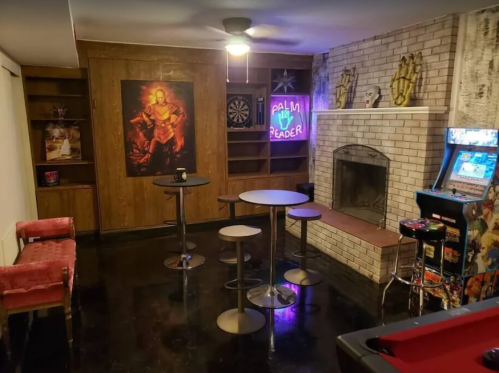A cozy game room featuring a dartboard, arcade machine, bar tables, and a neon sign, with a fireplace and retro decor.