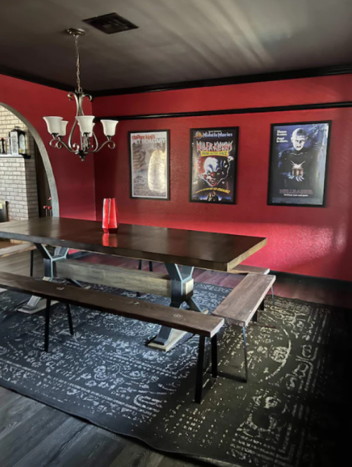 A dining room with a red wall, a wooden table, benches, and framed horror movie posters.