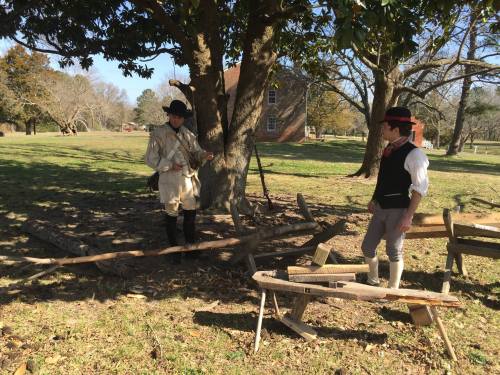 Two men in historical attire stand by a tree, engaged in a discussion near wooden tools in a grassy area.