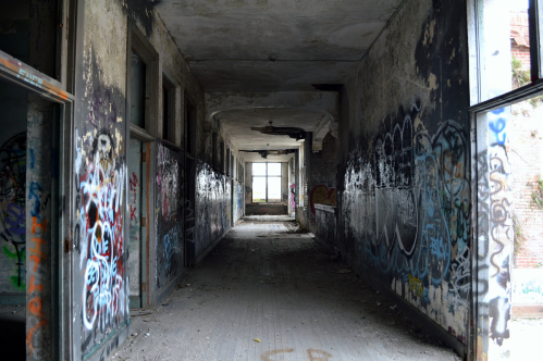 A dimly lit, abandoned hallway with graffiti-covered walls and a window at the far end. Dust and debris on the floor.