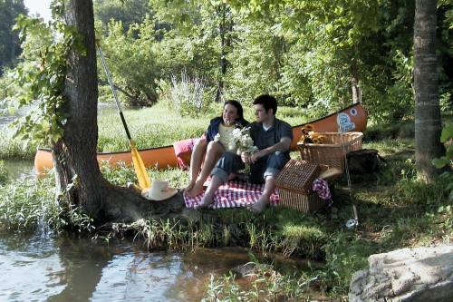 A couple sits by a riverbank, enjoying a picnic with a canoe nearby, surrounded by greenery and flowers.