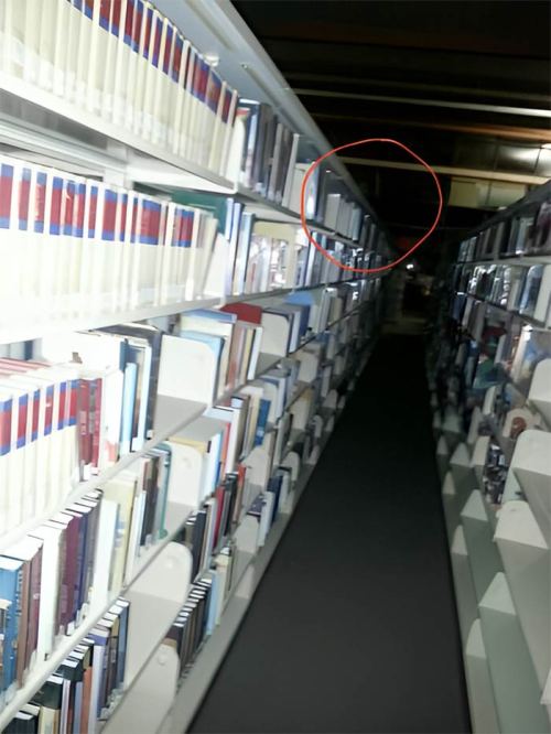 A dimly lit library aisle with bookshelves on either side and a highlighted section at the top of a shelf.