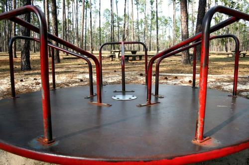 A rusty metal merry-go-round in a wooded park, surrounded by tall trees and picnic tables in the background.