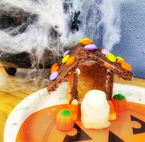 A Halloween-themed gingerbread house decorated with candy, surrounded by spider web decorations on a festive plate.