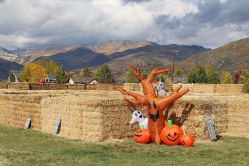 A festive scene with hay bales, inflatable Halloween decorations, and pumpkins against a backdrop of mountains and clouds.