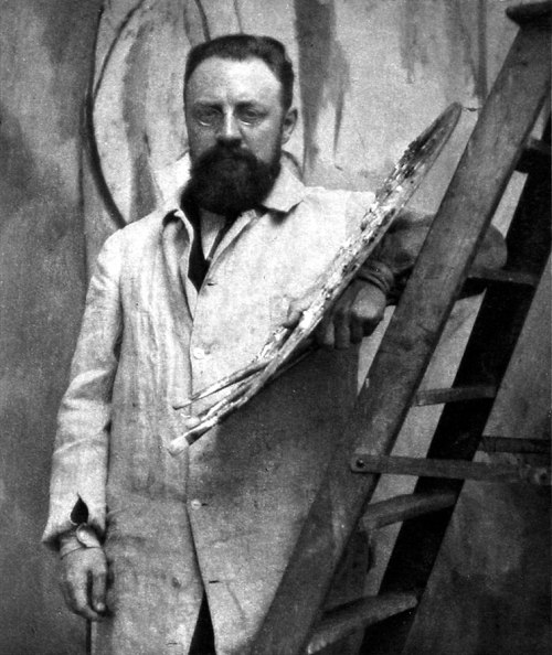 A bearded man in a white coat stands beside a wooden ladder, holding a palette in a studio setting.