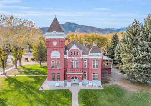 Historic red brick building with a clock tower, surrounded by trees and mountains in the background.