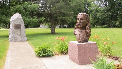 A bronze bust of a man with long hair stands beside a stone monument in a grassy park with trees and flowers.